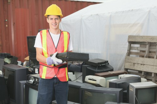 A young man wearing a yellow safety helmet, a high-visibility safety vest with reflective orange and yellow stripes, and work gloves, standing outdoors on a paved surface beside a collection of discarded electronic items including old computer monitors and printers. Behind him is a white temporary shelter or tent, and to the right, there is a weathered wooden pallet. The area appears to be part of a waste disposal or recycling yard, with a red metal fence visible in the background. The man is smiling and holding a black electronic device, suggesting a professional approach to rubbish collection and disposal, in line with services offered by Commercial Waste Removal Holborn in the central London area, close to Holborn postcode areas. The scene captures the process of managing electronic waste as part of rubbish removal activities, situated in an urban environment typical of the Holborn district.