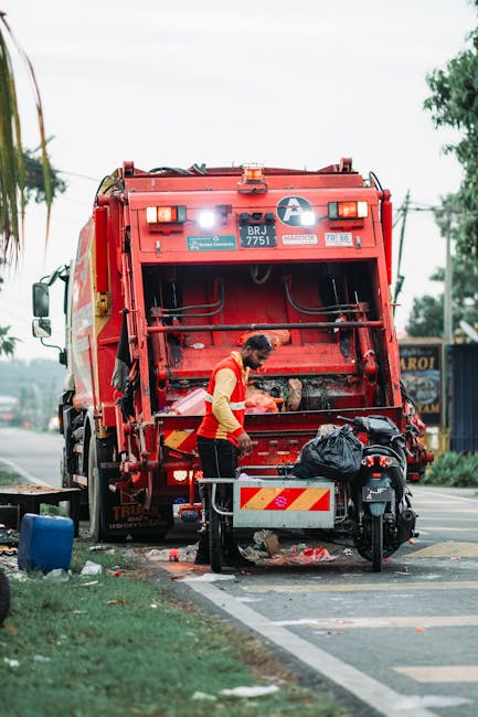 A large pile of mixed waste materials is stacked on a pavement or driveway in an outdoor setting. The heap includes cardboard boxes, plastic containers, and assorted refuse, with some items appearing